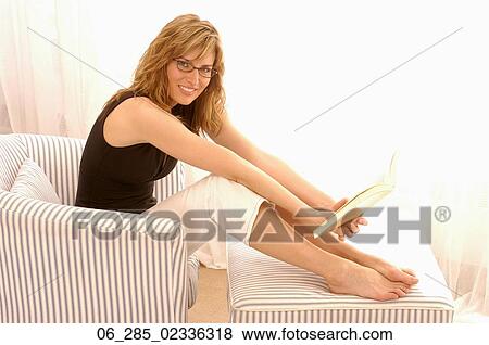 Full length side view blonde woman in glasses leaning forward from an armchair holding a book with her feet up looking at camera smiling View Large Photo Image Stock Photo - Full length side view blonde woman in glasses leaning forward from an armchair holding a book with her feet up looking at camera smiling. Fotosearch