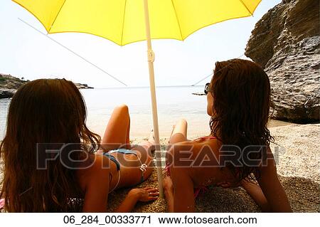 Stock Image - Half length rear view two women long brown hair lying side by side on a beach sunbathing under a yellow parasol looking out to sea. Fotosearch