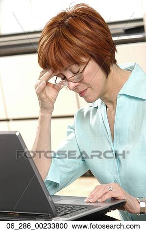 Stock Image - Half length side view woman in glasses sitting at a laptop leaning on her elbow with her eyes closed. Fotosearch