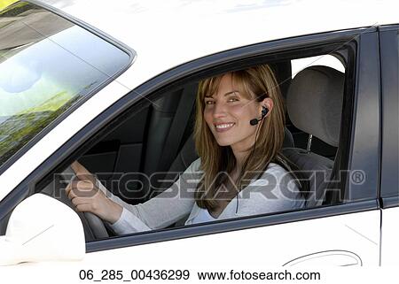 Stock Photo - Head and shoulders side view woman in drivers seat wearing an earphone looking at camera smiling. Fotosearch