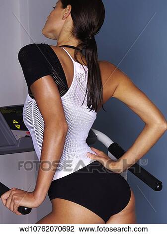 Stock Image - View from behind of female with long brunette hair wearing black leotard under white string vest standing on step machine hand on hip.. Fotosearch