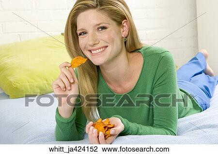 Stock Image -  Blonde woman in her 20's lying on a bed eats potato chips.. Fotosearch