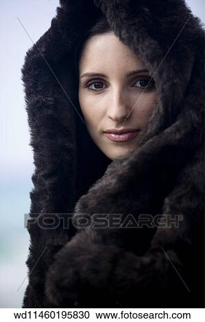 Stock Image - Female with brunette hair off face wearing black fur coat and hood looking straight to camera unsmiling.. Fotosearch