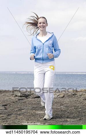 Female with long blonde hair off face wearing blue hooded top and white t shirt with white tracksuit trousers and trainers running towards camera with sea in background smiling showing teeth. View Large Photo Image Picture - Female with long blonde hair off face wearing blue hooded top and white t shirt with white tracksuit trousers and trainers running towards camera with sea in background smiling showing teeth.. Fotosearch