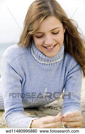 Stock Image - Female with long blonde hair wearing pale blue poloneck jumper lying forward on sand leaning up on elbows looking down at hands smiling showing teeth. . Fotosearch