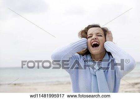 Stock Photo - Female with long blonde hair wearing pale blue zip up jumper and matching scarf standing on beach with sea in background holding hands to ears head tilted back eyes closed mouth open showing teeth. . Fotosearch