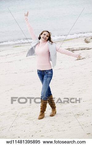 Stock Photography - Female with long blonde hair wearing pink poloneck jumper and blue jeans tucked into tan suede boots and pale blue sleeveless jacket walking on sand arms outstretched looking up smiling showing teeth.. Fotosearch