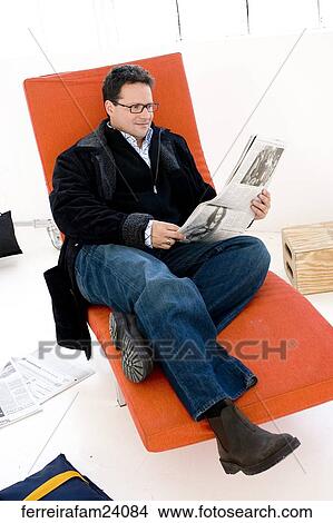 Man in his 30's wearing glasses reading a newspaper sitting on a red chair. View Large Photo Image Picture - Man in his 30's wearing glasses reading a newspaper sitting on a red chair.. Fotosearch