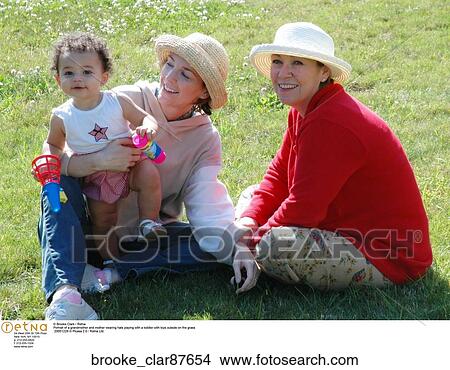 Picture - Portrait of a grandmother and mother wearing hats playing with a toddler with toys outside on the grass. Fotosearch