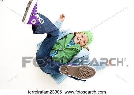 Stock Photography -  Portrait of a young girl lying on a white backdrop wearing a blue coat and green hat and purple sneakers. Fotosearch