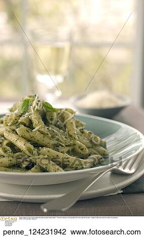 Stock Image -  Still life image of a bowl of penne pasta with pesto and green spices visible. The bowl is resting on a serving plate with a fork underneth the bowl. A glass of water and a bowl are visible in the backdrop.. Fotosearch