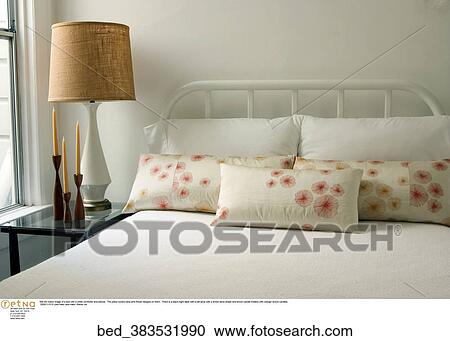 Stock Image -  Still life indoor image of a bed with a white comforter and pillows. The pillow covers have pink flower designs on them. There is a black night table with a tall lamp with a brown lamp shade and brown candle holders with orange/ brown candles.. Fotosearch