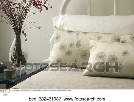 Still life indoor image of a bed with a white comforter and pillows. The pillow covers have faint flower designs on them. There is a black side table with a glass vase holding flowers next to the bed. View Large Photo Image Stock Photo - Still life indoor image of a bed with a white comforter and pillows. The pillow covers have faint flower designs on them. There is a black side table with a glass vase holding flowers next to the bed.. Fotosearch