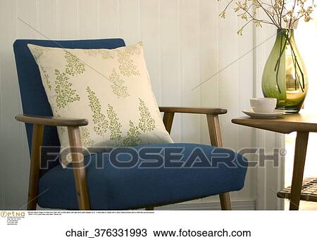 Stock Image -  Still life indoor image of a blue arm chair with a white pillow with light berry plant designs on it. A side table is visible with a vase holding flowers and a white tea cup and saucer.. Fotosearch