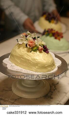 Still life indoor image of a cake covered with white frosting and topped with flower garnishes sitting on a silver serving platter. Other similar cakes are visible in the background. View Large Photo Image Stock Image - Still life indoor image of a cake covered with white frosting and topped with flower garnishes sitting on a silver serving platter. Other similar cakes are visible in the background.. Fotosearch