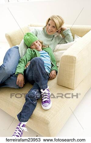 Stock Image -  Woman wering a green sweater and a young girl wearing a green hat and a green jacket sitting in a chair. The girl leans agains the woman. . Fotosearch