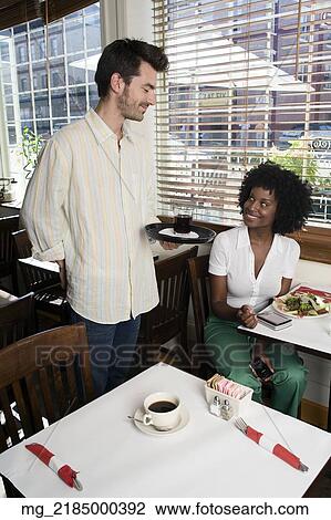 Stock Image - A waitor brings a young woman her 20's wearing a white short sleeve top and green pants a drink while she sits in a restaurant eating salmon and salad. September 1, 2006 credit - RD / Retna Digital. Fotosearch