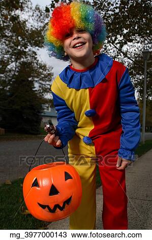 Stock Image - A young boy on the sidewalk on the quest for more halloween candy. He is dressed in a yellow, red and blue clown costume accompanied with a rainbow wig. He is holding a pumpkin filled with candy. © Retna Digital / Retna Ltd.. Fotosearch