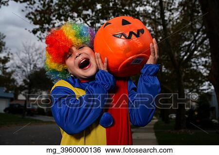 Stock Photograph - A young boy on the sidewalk on the quest for more halloween candy. He is dressed in a yellow, red and blue clown costume accompanied with a rainbow wig. He is holding a pumpkin filled with candy. © Retna Digital / Retna Ltd.. Fotosearch