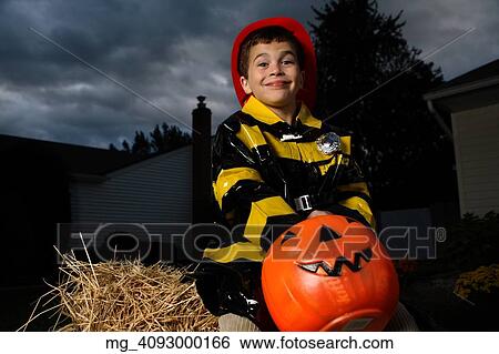 Stock Photograph - A young boy out on halloween. He is wearing a fireman costume, equipped with hat & coat. He's outside sitting on a haystack with his pumpkin filled with candy. © Retna Digital / Retna Ltd.. Fotosearch
