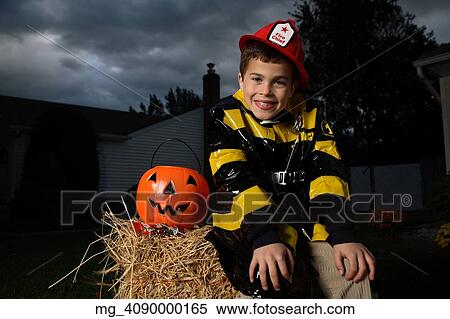 Stock Photography - A young boy out on halloween. He is wearing a fireman costume, equipped with hat & coat. He's outside sitting on a haystack with his pumpkin filled with candy. © Retna Digital / Retna Ltd.. Fotosearch