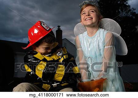 A young girl & boy out on halloween. He is wearing a fireman costume, equipped with hat & coat. The Girl is dressed as a fairy princess in a light blue gown, wings and a halo. They are outside sitting on a haystack playing. © Retna Digital / Retna Lt View Large Photo Image Stock Image - A young girl & boy out on halloween. He is wearing a fireman costume, equipped with hat & coat. The Girl is dressed as a fairy princess in a light blue gown, wings and a halo. They are outside sitting on a haystack playing. © Retna Digital / Retna Lt. Fotosearch