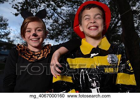 Stock Image - A young girl & boy out on halloween. She is dressed as a cat in a black unitard and tights with a animal print fur trim collar. She uses makeup to a cat noses and whiskers and costume ears. The Boy is wearing a fireman costume, equipped with hat & coat. Fotosearch