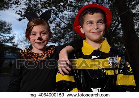Stock Photo - A young girl & boy out on halloween. She is dressed as a cat in a black unitard and tights with a animal print fur trim collar. She uses makeup to a cat noses and whiskers and costume ears. The Boy is wearing a fireman costume, equipped with hat & coat. Fotosearch