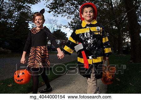 Picture - A young girl & boy walking down the sidewalk on a quest for more halloween candy. She is dressed as a cat in a black unitard and tights with a animal print fur trim collar and skirt. She uses makeup to a cat noses and whiskers and costume ears. The Boy. Fotosearch