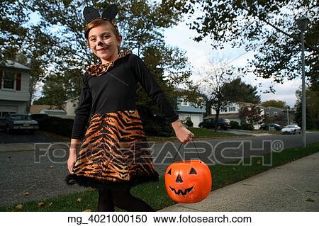 Stock Image - A young girl on the sidewalk on the quest for more halloween candy. She is dressed as a cat in a black unitard and tights with a animal print fur trim collar and skirt. She uses makeup to a cat noses and whiskers and costume ears. She is holding a pump. Fotosearch