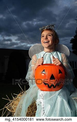 Stock Image - A young girl out on halloween. She is dressed as a fairy princess in a light blue gown, wings and a halo. She is sitting outside on a haystack with a pumpkin filled with candy. © Retna Digital / Retna Ltd.. Fotosearch