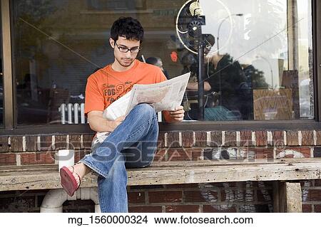 A young man in glasses, orange shirt and blue jeans, and flip flops reads a newspaper outside a shop while enjoying a drink. September 1, 2006 credit - RD / Retna Digital View Large Photo Image Picture - A young man in glasses, orange shirt and blue jeans, and flip flops reads a newspaper outside a shop while enjoying a drink. September 1, 2006 credit - RD / Retna Digital. Fotosearch