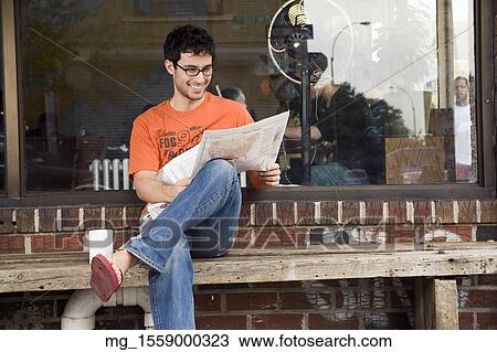 A young man in glasses, orange shirt and blue jeans, and flip flops reads a newspaper outside a shop while enjoying a drink. September 1, 2006 credit - RD / Retna Digital View Large Photo Image Stock Image - A young man in glasses, orange shirt and blue jeans, and flip flops reads a newspaper outside a shop while enjoying a drink. September 1, 2006 credit - RD / Retna Digital. Fotosearch
