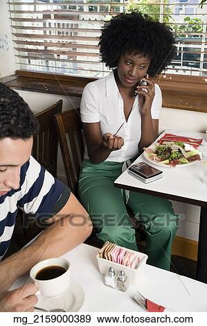 A young woman her 20's wearing in a white short sleeve top and green pants talks on her cell phone while sitting in a restaurant eating salmon and salad while using her Personal Digital Assistant as a young man sits drinking coffee in the foreground. Sep View Large Photo Image Stock Photo - A young woman her 20's wearing in a white short sleeve top and green pants talks on her cell phone while sitting in a restaurant eating salmon and salad while using her Personal Digital Assistant as a young man sits drinking coffee in the foreground. Sep. Fotosearch