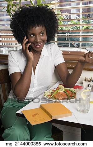 Stock Photography - A young woman her 20's wearing in a white short sleeve top and green pants talks on her cell phone while sitting in a restaurant eating salmon and salad while reading a book. September 1, 2006 credit - RD / Retna Digital. Fotosearch