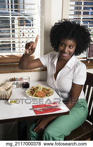 Stock Photo - A young woman in her 20's wearing a white short sleeve top and green pants sits in a restaurant and gestures to get the waitor's attention or ask for the check. September 1, 2006 credit - RD / Retna Digital. Fotosearch