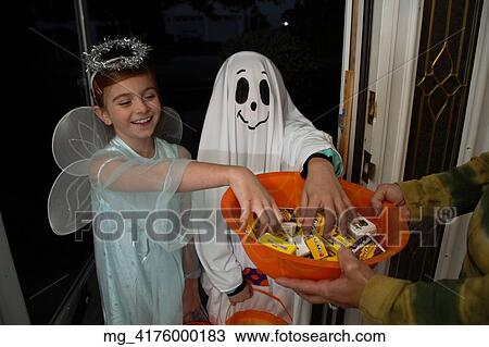 Stock Image - Children out on halloween. The girl is dressed as a fairy princess in a light blue gown, wings and a halo. The other child is dressed as a ghost. They are in a doorway getting thier candy treats. © Retna Digital / Retna Ltd.. Fotosearch