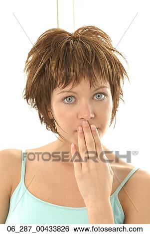 Stock Photograph - Head and shoulders front view young woman looking at the camera wide eyed with hand over her mouth. Fotosearch