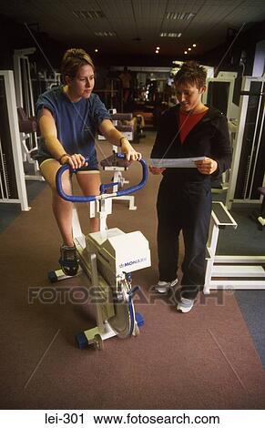 Stock Image - Woman on Exercise Bike and Instructor UK. Fotosearch