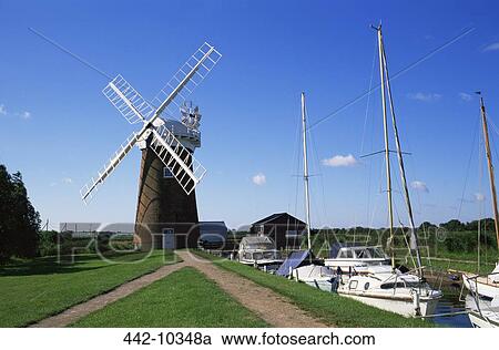 Stock Image - Boats moored near a traditional windmill, Horsey Windpump, Horsey, Norfolk Broads, Norfolk, England. Fotosearch