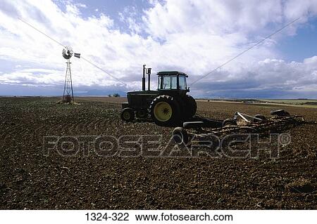 Stock Image - Farm tractor in a field with an irrigation well and a windmill in the background, Dove Creek, Colorado, USA. Fotosearch