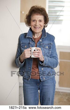 Stock Image - Senior woman standing in the kitchen and drinking coffee. Fotosearch