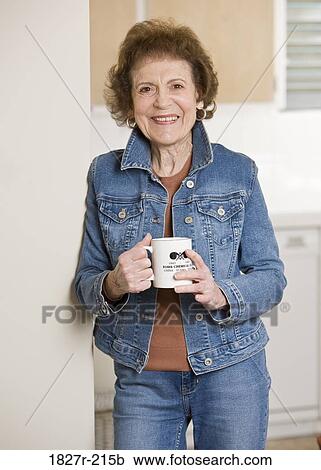 Stock Image - Senior woman standing in the kitchen and drinking coffee. Fotosearch