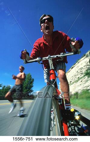 Stock Image - Low angle view of a young man riding a cycle with another young man running behind him. Fotosearch