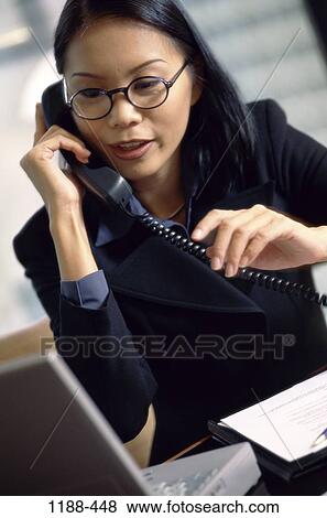 Stock Photo - desk, woman, telephone, talking, Asian, eyeglasses, laptop. Fotosearch