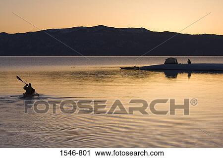 Stock Image - Silhouette of a man kayaking in a lake with a woman near a tent in the background, Lake Tahoe, California, USA. Fotosearch