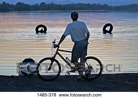 Rear view of a man standing with his bicycle at the lakeside with two people swimming in the lake View Large Photo Image Stock Photo - Rear view of a man standing with his bicycle at the lakeside with two people swimming in the lake. Fotosearch