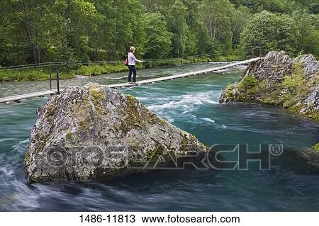 Stock Image - Woman crossing a river by a footbridge, Flamsdalen Valley River, Flam, Aurlandsfjord, Sogn Og Fjordane, Norway. Fotosearch