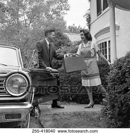 Stock Image - Housewife passing suitcase to husband in front of house. Fotosearch