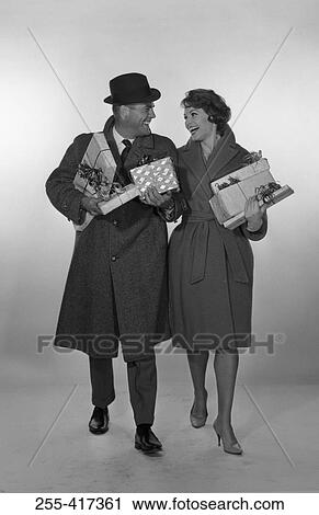 Stock Image - Mid adult couple laughing, looking at each other and holding gifts, studio shot. Fotosearch